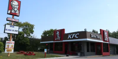 A KFC fast-food restaurant with a red and white exterior, a prominent Colonel Sanders sign, and a drive-thru, set against a clear blue sky with picnic tables and trees nearby.