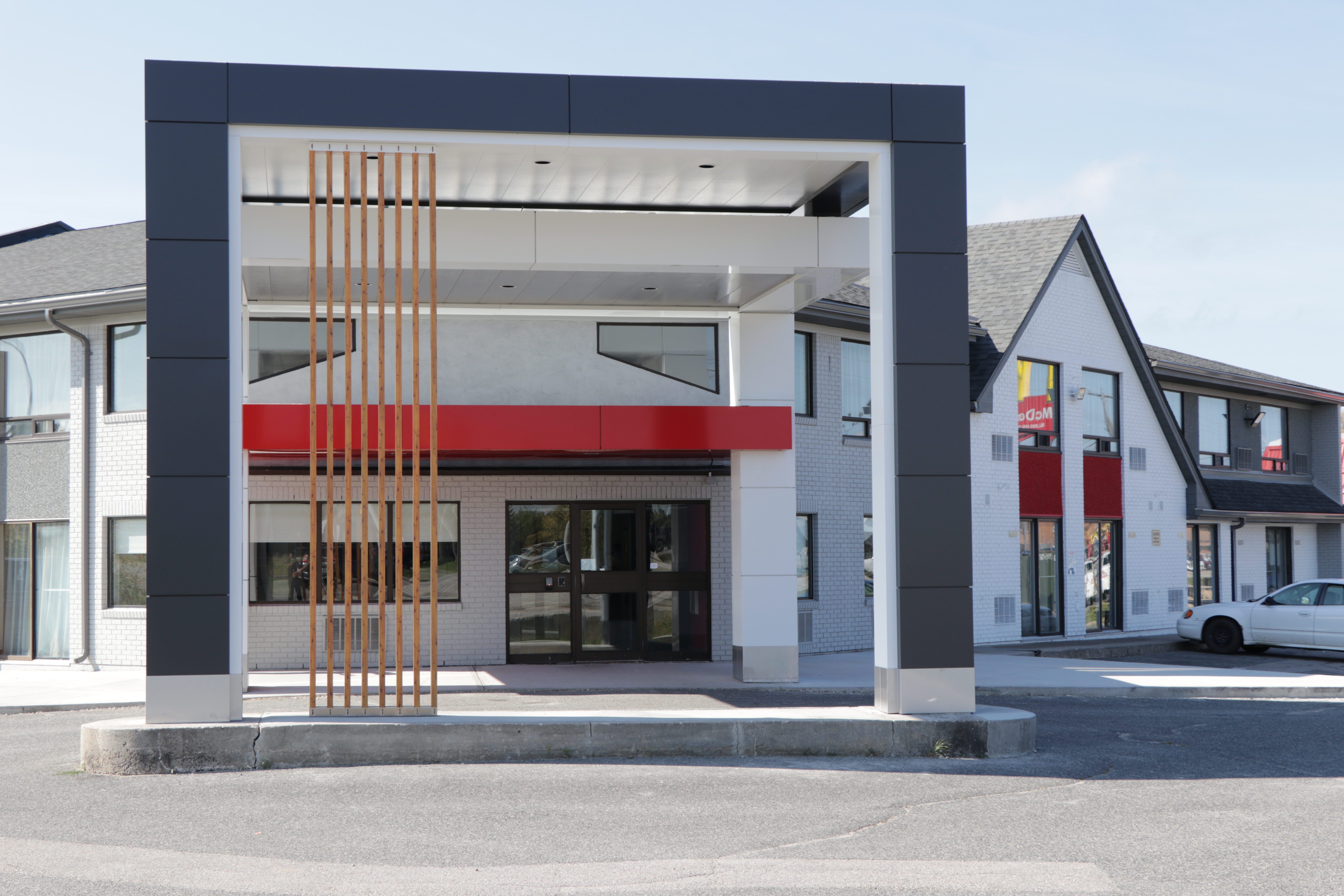 A modern hotel building with a glass-paneled entrance under a canopy, supported by columns, featuring a lodgepole timber facade, parking lot, and urban setting under a clear sky.