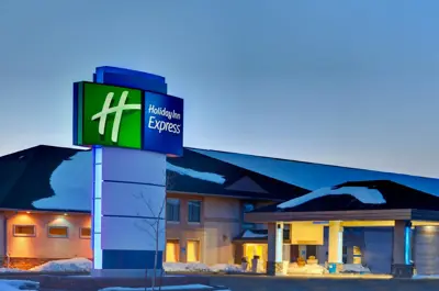 A modern Holiday Inn hotel exterior with a well-lit entrance, featuring the brand’s green and yellow sign, surrounded by a snowy parking lot and landscaped bushes, under a clear evening sky.