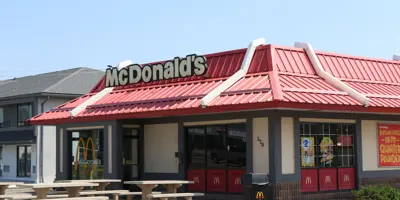 A McDonald's restaurant with a red roof and golden arches sign, featuring a drive-thru, outdoor seating with picnic tables, and a menu board, set in an urban parking lot.
