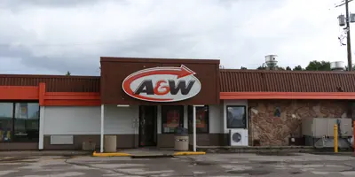 An A&W restaurant with a red and white sign featuring the brand logo, set against a cloudy sky with a cracked parking lot and a stone facade.