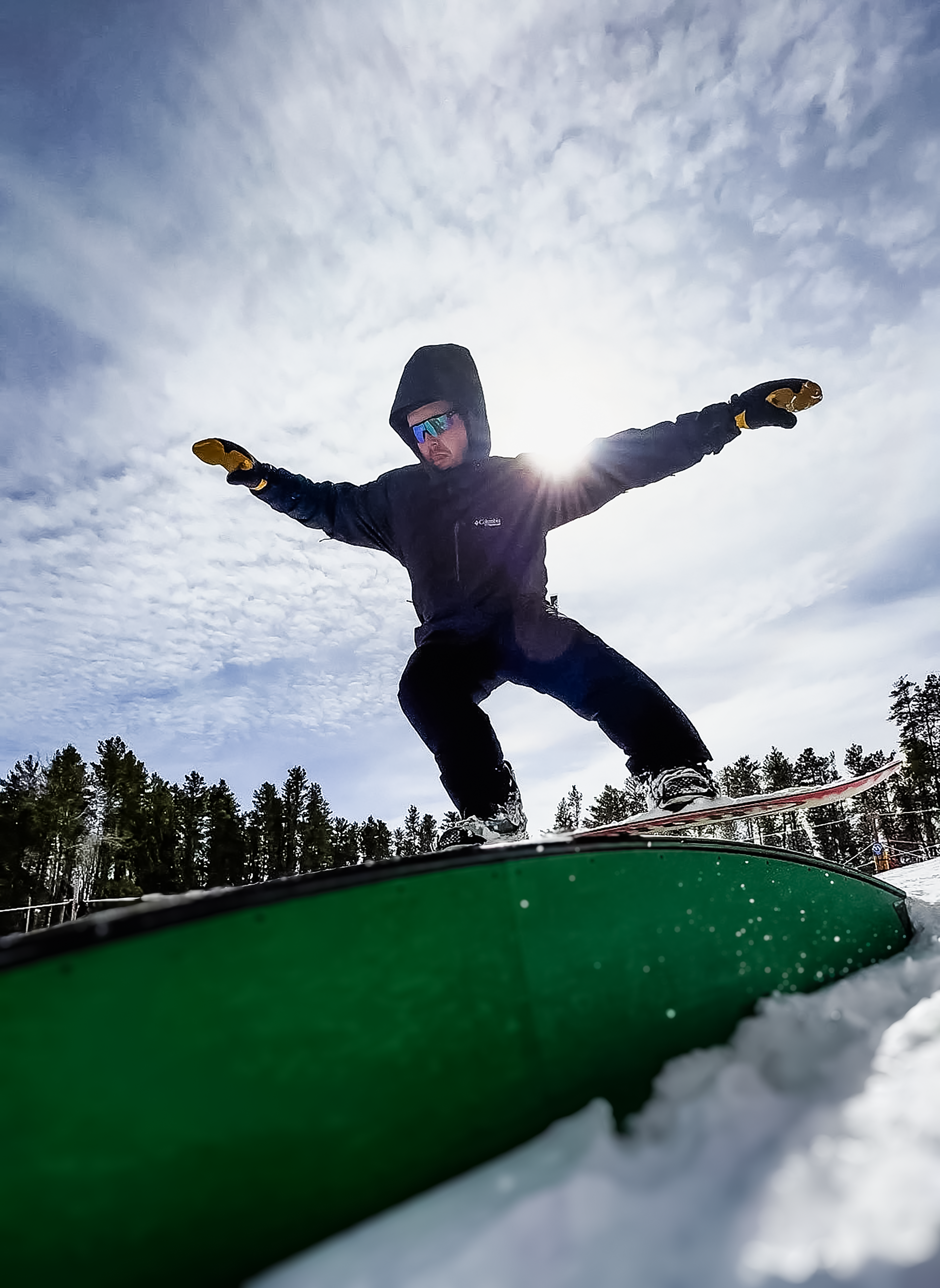 A snowboarder in mid-air performing a trick on a snowy slope, wearing a jacket, gloves, and goggles, with a sunny sky, forested landscape, and fresh powder in the background.