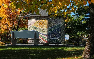 A colorful mosaic-covered structure in a park, surrounded by trees with vibrant autumn leaves in shades of yellow and orange, under a sunny sky.