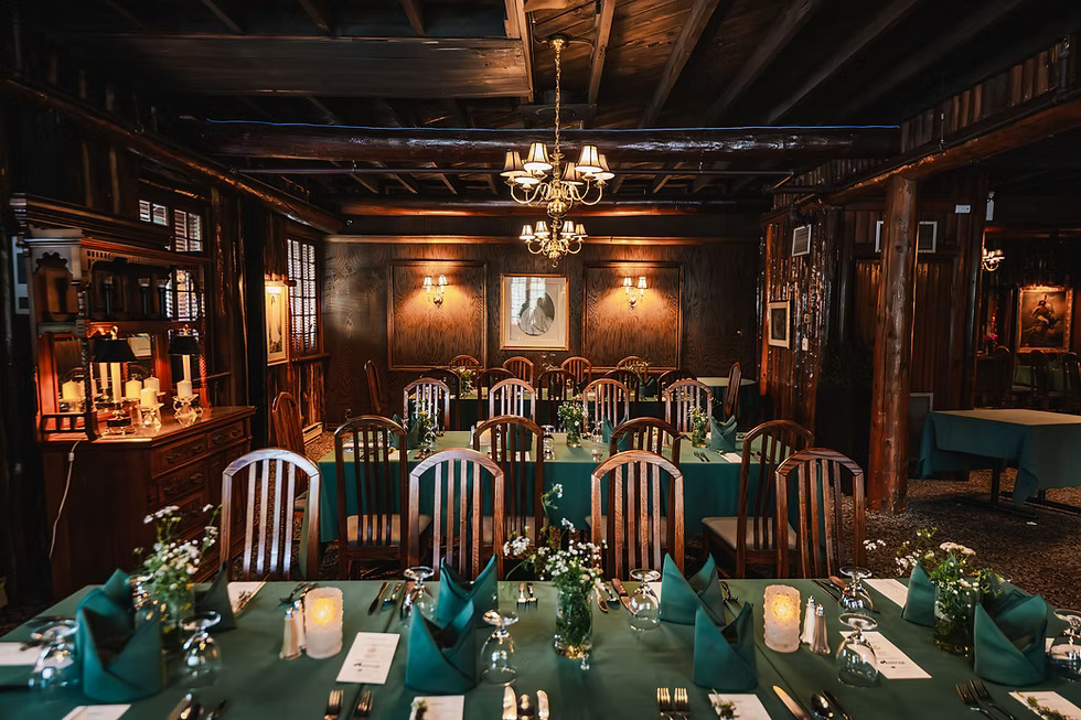 An elegant dining room with wooden beams, a chandelier, and a long table set with napkins and candles, exuding a warm, upscale ambiance for a formal event.