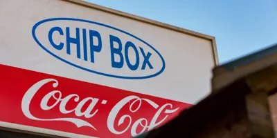 A signboard advertisement featuring a Coca-Cola logo on a building roof edge, promoting chips and fries in a red, white, and blue container, set against a clear blue sky.