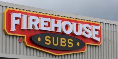  A Firehouse Subs restaurant exterior with a red, yellow, and white sign featuring a maple leaf emblem, indicating a Canadian location, under a canopy in an urban setting.