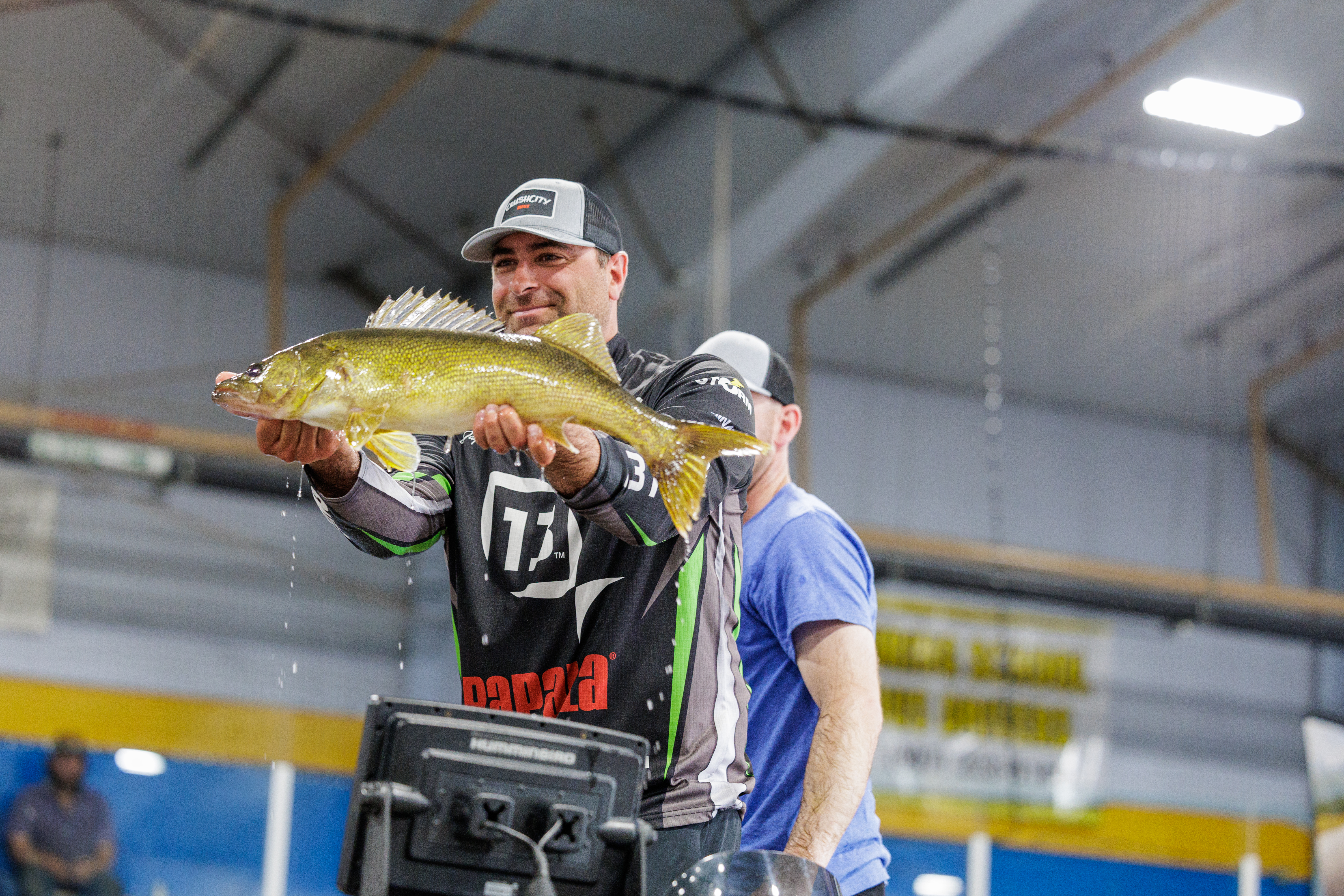 An angler holding up a large walleye indoors during a fishing competition, water dripping from the fish