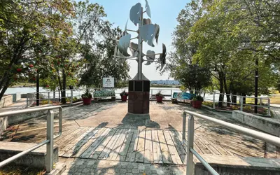 A modern metal sculpture in an outdoor park with trees, a boardwalk, and a lake waterfront, featuring a bench and railing under a sunny sky.