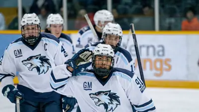 Hockey player in a white and blue dog-logo jersey celebrates on the ice while teammates follow behind during a game