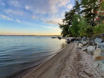 A sandy beach with calm water and scattered rocks, with the sky glowing in shades of blue and orange at sunset