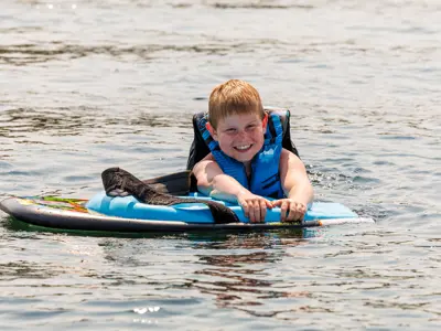 Smiling young boy in a life jacket riding a water board on a lake on a sunny day