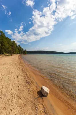 A long stretch of sandy beach beside clear lake water, bordered by trees under a bright blue sky with scattered clouds
